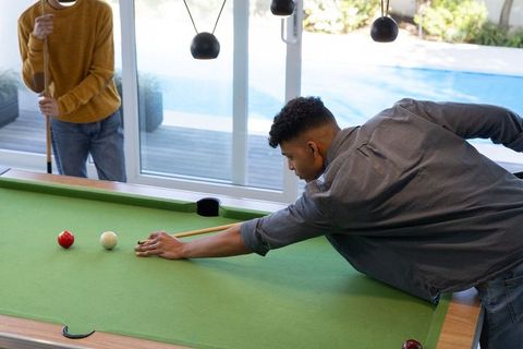 Young men playing pool in modern home game room leaning over green felt table