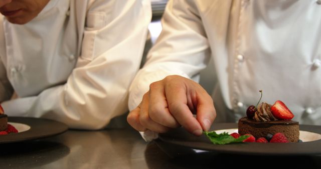 Two chefs in white uniforms are carefully plating elegant chocolate desserts with raspberries and strawberries in a professional kitchen. This image can be used in materials related to culinary arts, gourmet cooking, restaurant promotions, chef training programs, or food photography.