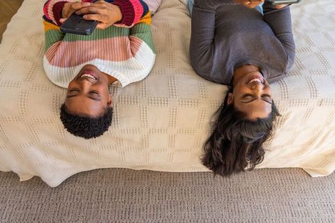Overhead portrait diverse female friends lying on bed smiling using smartphones