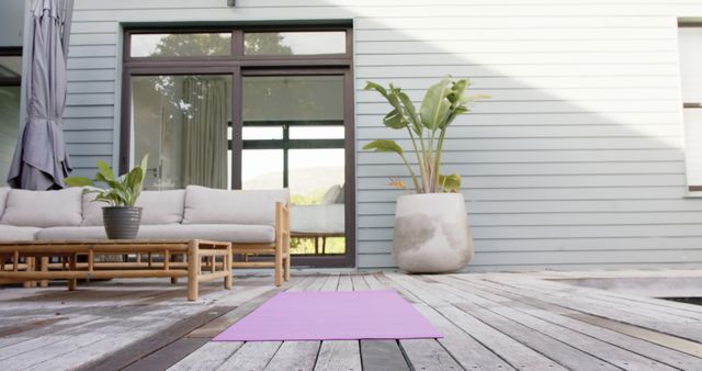 Image shows a purple yoga mat placed on a wooden deck outside a modern house with large windows. Nearby, there is a comfortable patio set and large indoor plant, creating a serene and inviting environment. Ideal for promoting wellness, relaxation, outdoor activities, or healthy lifestyle content.