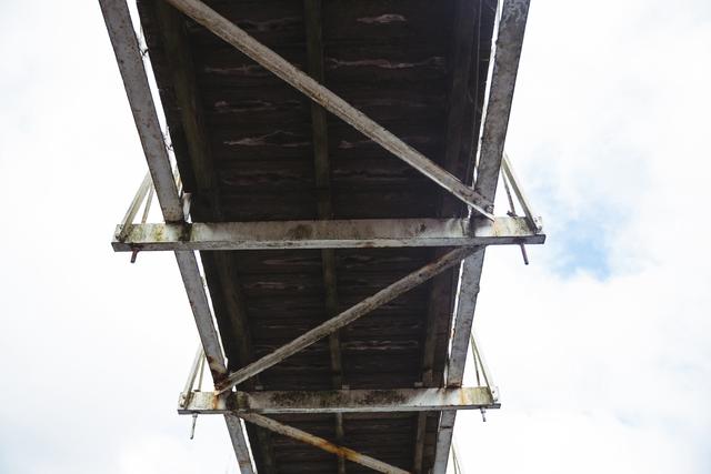 Low angle view of an old, rusty bridge showcasing its metal structure against a cloudy sky. Useful for illustrating themes of decay, industrial architecture, and urban infrastructure. Ideal for use in articles, presentations, or projects related to engineering, construction, and urban development.