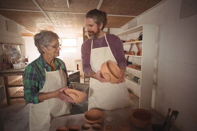 Senior woman and young man examining clay pots in a pottery workshop. They are wearing aprons and surrounded by various pottery items. This image can be used for themes related to art, craftsmanship, learning, and intergenerational activities.