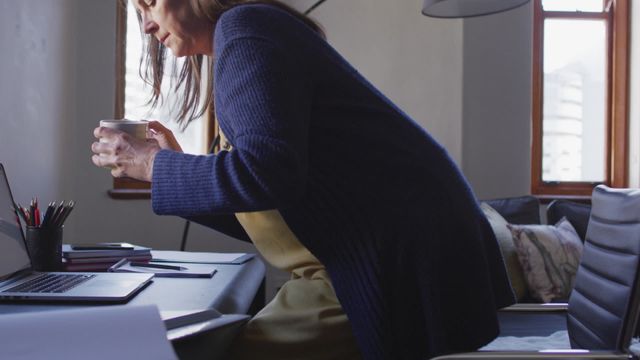 Woman sitting at a desk in a home setting, holding a cup of coffee. Ideal for concepts related to remote work, pandemic lockdowns, social distancing, and everyday lifestyle at home. Suitable for use in articles about work-life balance, working from home, or relaxation routines.