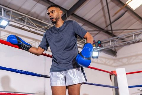 Male boxer leaning on ring ropes after sparring in indoor arena, wearing blue gloves