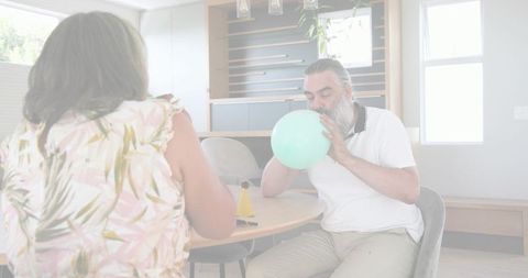 Senior man inflating pale green balloon at dining table with woman in floral top