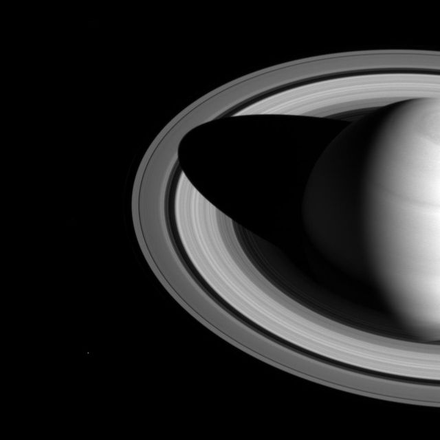 Stunning visual showing shadow of Saturn on rings, captured by Cassini spacecraft. Displays changing shadow length indicating the seasonal cycles on Saturn as it nears northern hemisphere solstice. Ideal for educational content, space-related articles, and science presentations. High-quality image showcases sunlit side of the rings with a clear view of Saturn's moon, Mimas, visible at lower left.