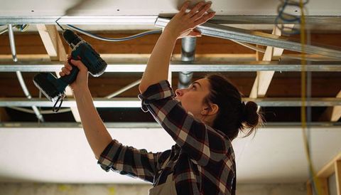 Woman drilling ceiling drywall with cordless drill securing metal track during renovation