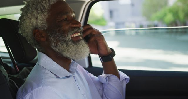 Senior man with gray beard and white hair smiling while having phone call in car during daytime. Useful for topics on senior mobility, technology use among elders, travel convenience, and communication.