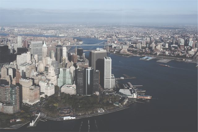 An aerial view showcases the iconic skyline of Lower Manhattan, highlighting the juxtaposition of modern skyscrapers and historic architecture. You can see parts of the Hudson River and East River framing the landscape, with the Brooklyn Bridge prominently in the background connecting Manhattan to Brooklyn. This image is ideal for urban infrastructure studies, articles about New York City, real estate promotions, and travel brochures emphasizing the allure of downtown Manhattan.