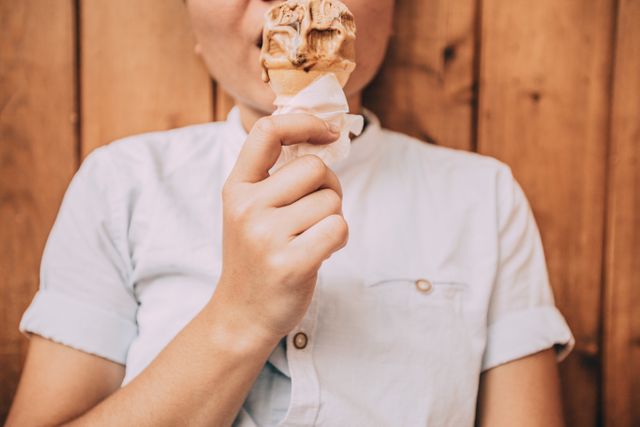 Young individual holding and consuming ice cream cone with mix of vanilla and chocolate flavors. Person wearing a light blue shirt, wooden background. Perfect for summer-themed promotions, dessert advertisements, leisure activities, young audience targeting.