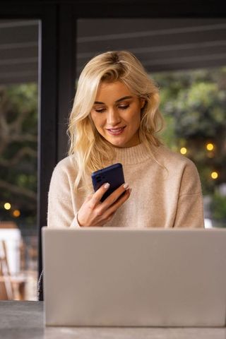 Blonde woman working remotely in cozy cafe wearing beige sweater using smartphone and laptop