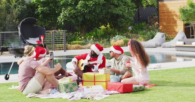 Group of friends celebrating Christmas outside. They are sitting on the grass, exchanging gifts, and enjoying music played on the guitar. They are all wearing festive Santa hats and looking engaged in the moment. The background features a swimming pool and greenery, indicating a relaxing and joyful atmosphere. This image can be used to depict holiday joy, outdoor celebrations, or festive gatherings.