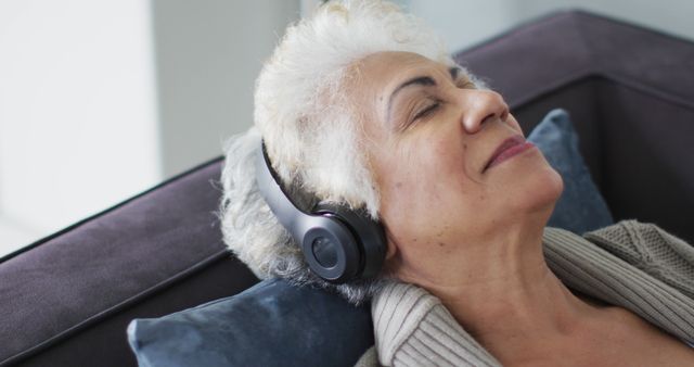 A senior woman is resting on a sofa, wearing headphones and appearing to enjoy listening to music. Her serene expression conveys relaxation and comfort, making this perfect for use in advertisements for music streaming services, health and wellness campaigns, senior living communities, and articles on relaxation or mental well-being.