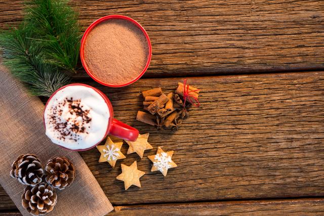 This festive scene features a cup of hot chocolate topped with whipped cream and cocoa powder, surrounded by cinnamon sticks, star-shaped cookies, pine cones, and a sprig of fir on a rustic wooden table. Ideal for holiday-themed advertisements, social media posts, greeting cards, or blog articles about Christmas, winter beverages, or cozy home decor.