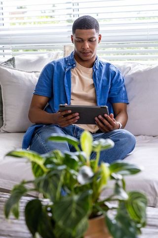 Young african american man relaxing cross-legged on sofa using tablet with indoor plant