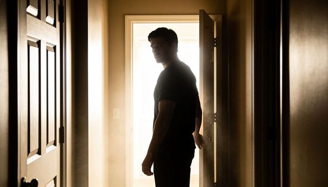 Silhouetted man standing in doorway turning head while holding doorknob backlit corridor