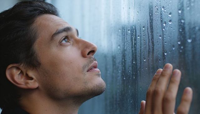 Contemplative man gazing through rain-streaked window pressing fingertips