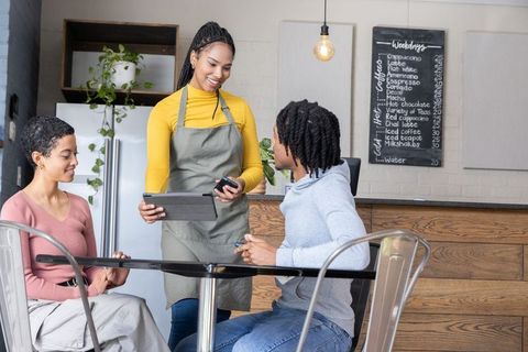 African american barista serving friends at cafe table using tablet pos for card payment