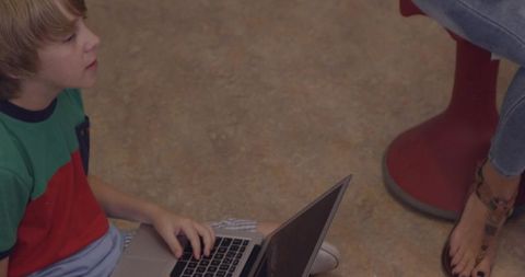 Young boy typing on laptop while sitting on floor beside adult, casual learning scene