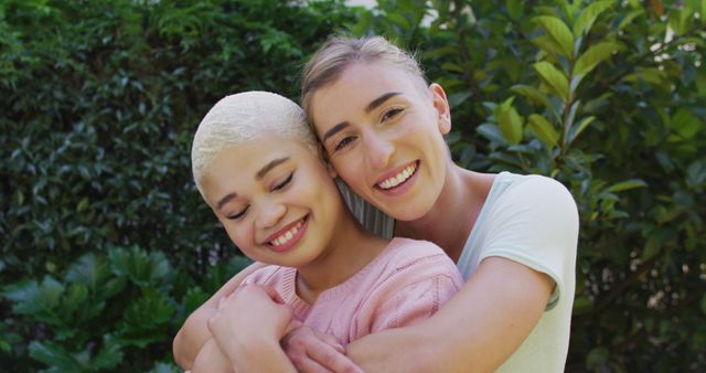 Two women are hugging and smiling outdoors, surrounded by lush greenery in a garden or park. This image can be used for promoting friendship, happiness, and diversity. It is ideal for use in lifestyle blogs, social media posts, and promotional materials related to wellness, nature, and relationships.