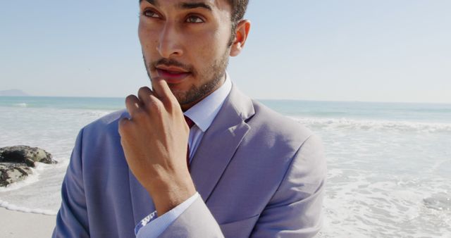 A young biracial man wearing suit stands on beach, touching his lips. He has short black hair, a beard, and dark eyes, looking thoughtful