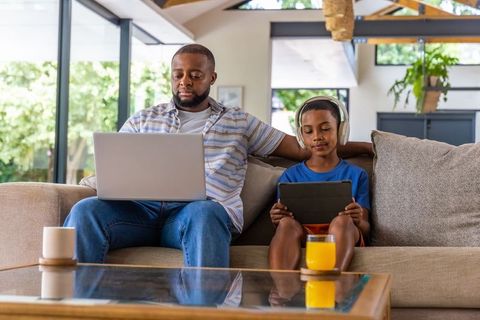 Black father working on laptop while son uses tablet on couch in modern living room