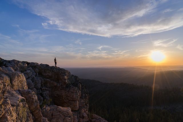Solitary person standing on edge of rocky cliff staring at sunset. Sunlight casts warm glow over expansive forest and horizon. Suitable for themes of exploration, adventure, natural beauty, solitude, serene landscapes, nature posters, and outdoor advertisements.