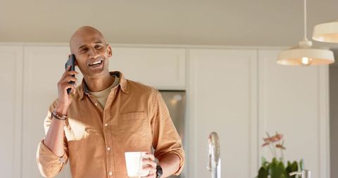 Mature african american man talking on phone holding coffee mug in modern kitchen