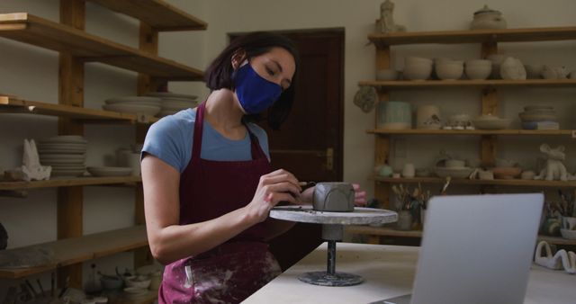 Female caucasian potter wearing face mask using ribbon tool to create designs on pot while having a image chat on laptop at pottery studio. social distancing in the pottery studio during coronavirus