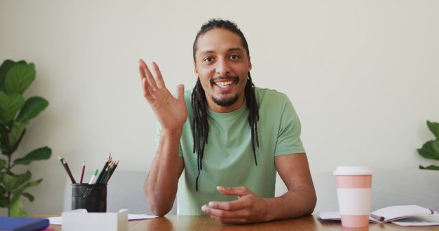 Man with dreadlocks sits at desk waving, offering a cheerful and welcoming gesture. Ideal for use in online communication, customer support imagery, working from home concepts, or friendly professional presentations. Background includes indoor plants and desk items like a pencil holder and coffee cup, indicating a relaxed, home office environment.