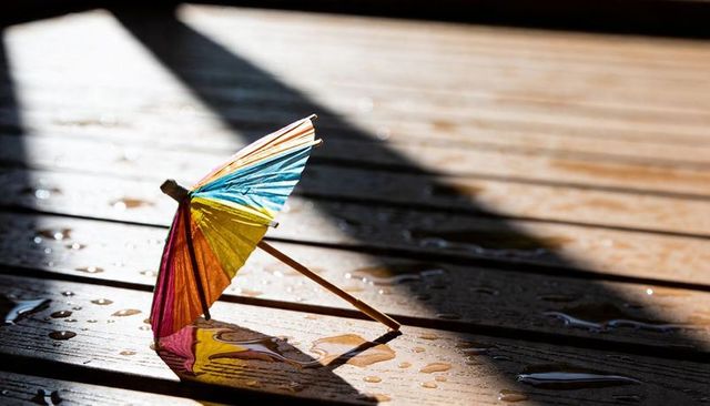 Colorful paper cocktail umbrella resting on wet wooden planks casting reflection