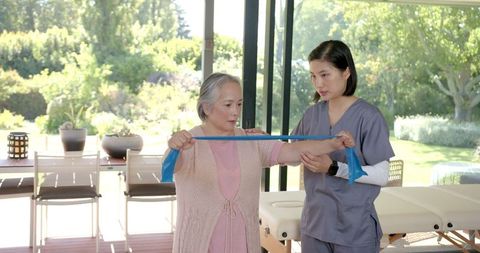 Senior Asian Woman Exercising with Resistance Band under Guidance of Physical Therapist
