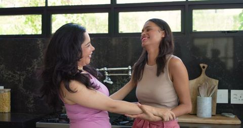 Intergenerational women laughing and holding forearms in sunlit kitchen, home bonding