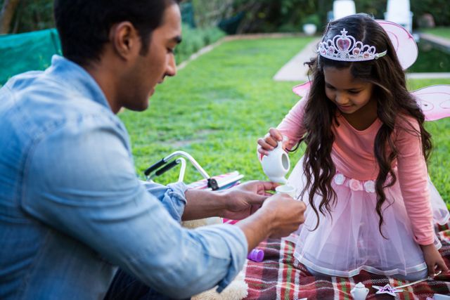 Father and daughter engaging in a toy tea party in a garden. The daughter is dressed in a fairy costume with wings and a tiara, pouring tea for her father. Ideal for use in family-oriented content, parenting blogs, advertisements promoting family activities, and articles about father-daughter relationships.