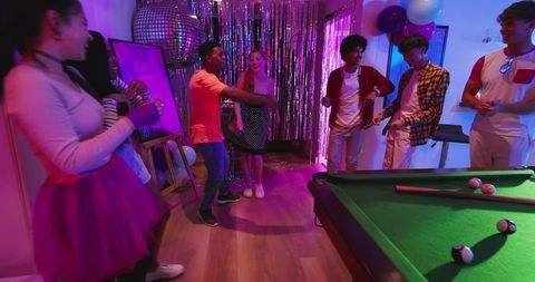 Multicultural Friends Dancing and Laughing Around Pool Table at Neon Disco House Party