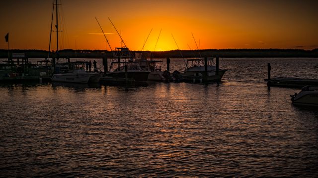 Serene sunset view at a marina with boats moored, casting silhouettes against a vivid orange horizon. Calm water reflects the tranquil twilight setting, ideal for illustrating concepts of peace, relaxation, boating lifestyles, or travel destinations.