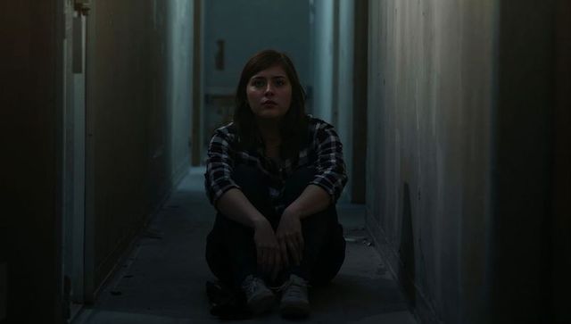 Solitary young woman sitting in narrow dim hallway clasping knees in moody low-key isolation