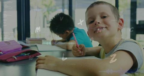 Smiling elementary boy writing at classroom desk while classmate studies in sunlit schoolroom