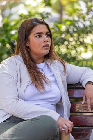 Young woman sitting on park bench holding coffee cup looking thoughtful in sunlit greenery