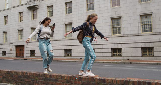 Two young women, dressed casually in jeans, walk on the ledge of a sidewalk wall in a city area. One is using her phone while the other extends her arms for balance. They appear to be enjoying a fun moment together. This image can be used to convey themes of friendship, adventure, and urban lifestyle.