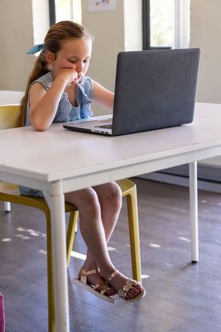 Bored school-aged girl using laptop at home study desk in sunlit minimalist room