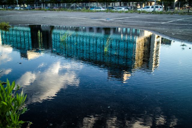 Reflection of a modern multi-story building in a puddle located in an empty parking lot. White clouds are reflected in the water along with the building, creating a serene and interesting visual effect. The image can be used for urban planning presentations, architecture portfolios, environmental awareness campaigns, and artistic projects emphasizing reflection and urban life.