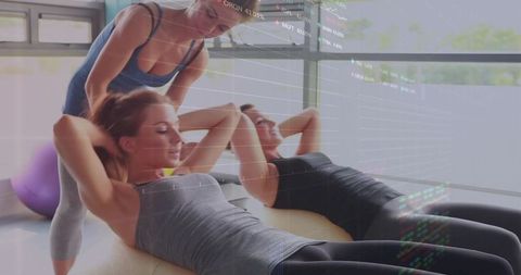 Female trainer guiding women through core crunches on mats with stability ball in sunlit gym studio