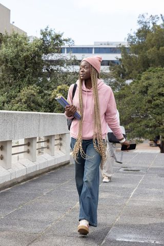 Female student walking on university campus bridge wearing pink hoodie holding notebooks