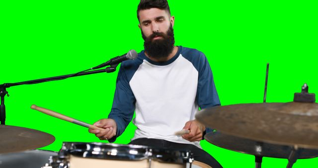 Bearded man in casual attire playing drums with a microphone nearby on green screen. This image works well for music-related projects, promotions for bands or solo musicians, and digital visual effects production mix-ins.