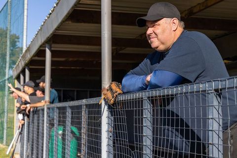 Veteran baseball coach leaning on dugout railing with worn glove, teammates watching