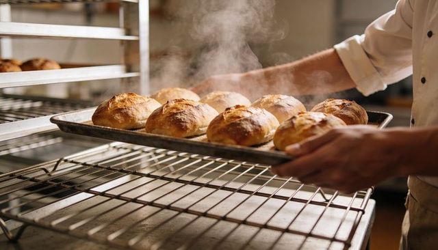 Baker placing tray of steaming golden rolls on cooling rack in artisanal bakery kitchen