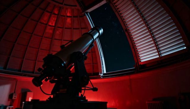 Refractor telescope silhouette inside red-lit observatory dome under starry night sky