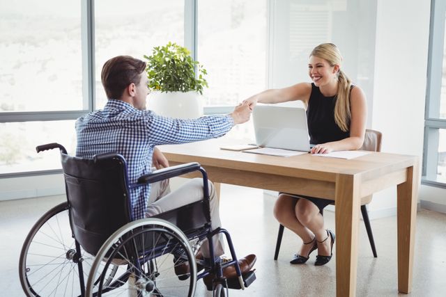 Two executives, one in a wheelchair, are shaking hands across a desk in a modern office. This image can be used to represent diversity and inclusion in the workplace, successful business negotiations, professional collaboration, and corporate partnerships.