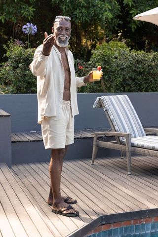 Mature african man smiling with cocktail at poolside in linen shirt and shorts, summer leisure